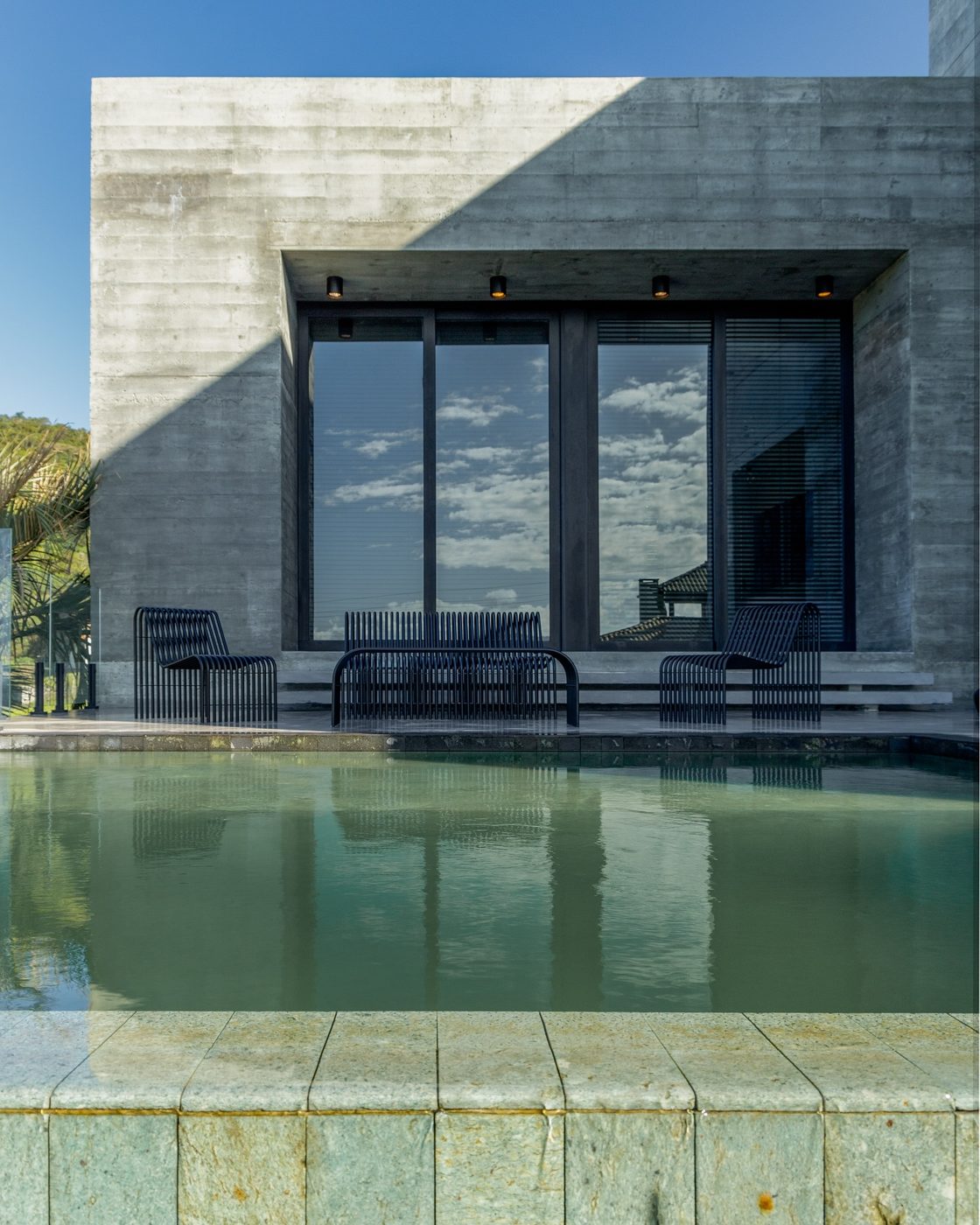 Casa de concreto aparente refletida na piscina, com céu azul