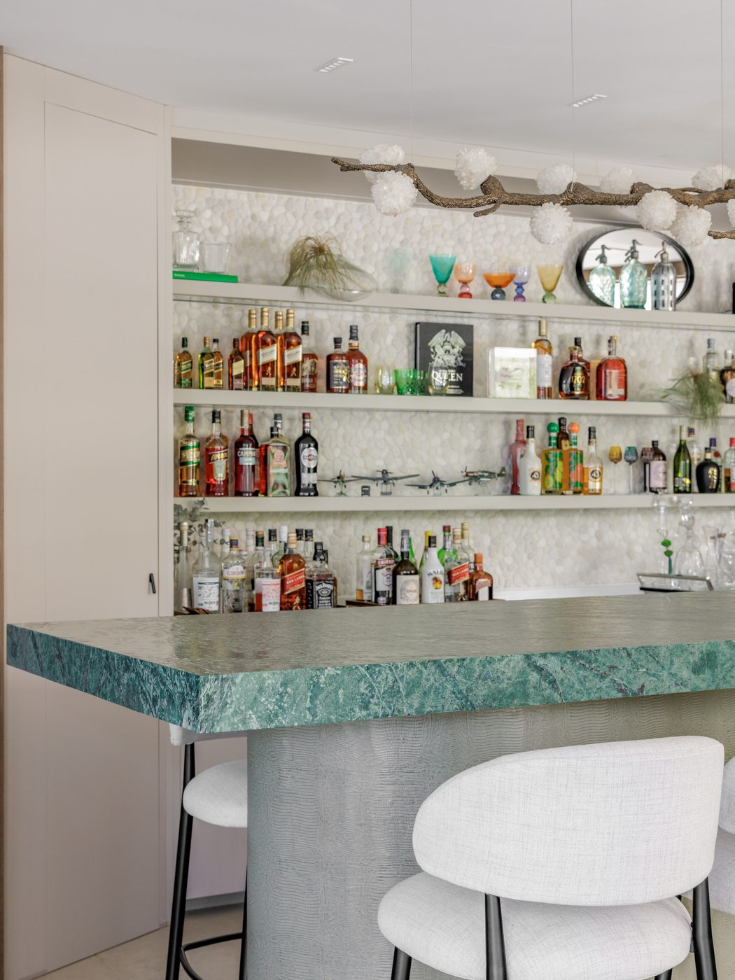 Bancada de bar em mármore verde com banquetas brancas e prateleiras de bebidas